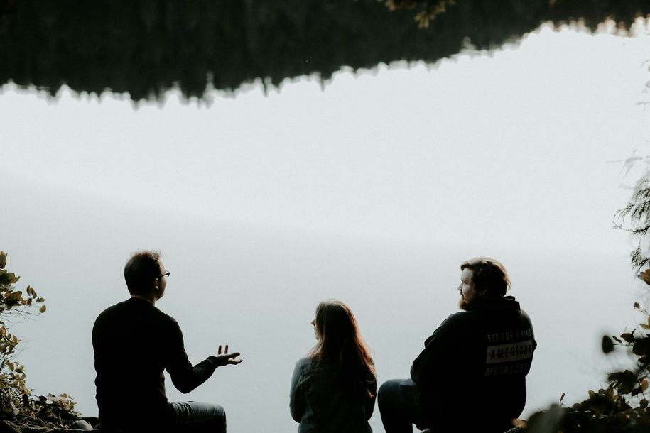 A group of people are sitting on the edge of a lake talking to each other.