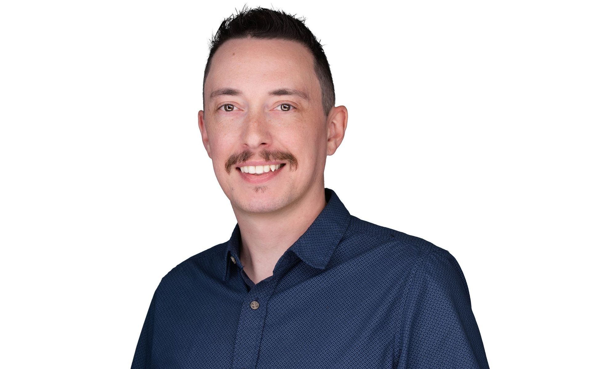 Man with mustache smiling, wearing a blue patterned button-up shirt against a white background.