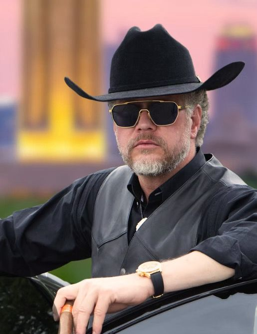 Man in cowboy hat and sunglasses leaning on car.