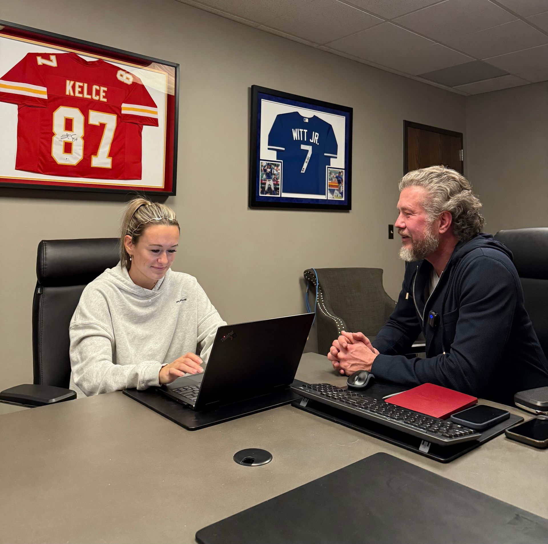 Woman and man seated at a desk, looking at a laptop. A football jersey and framed photo hang on the wall.