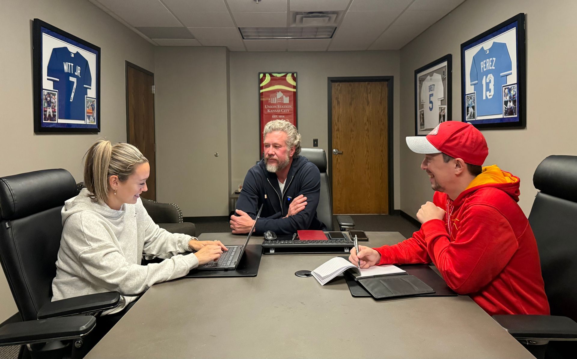 Three people sit around a table in an office. One works on a laptop, another takes notes. The middle person has arms crossed.