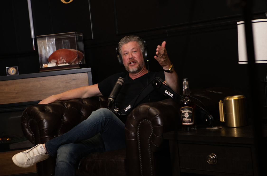 Man seated on a leather couch, gesturing while speaking into a microphone. Dark room with a liquor bottle on a side table.