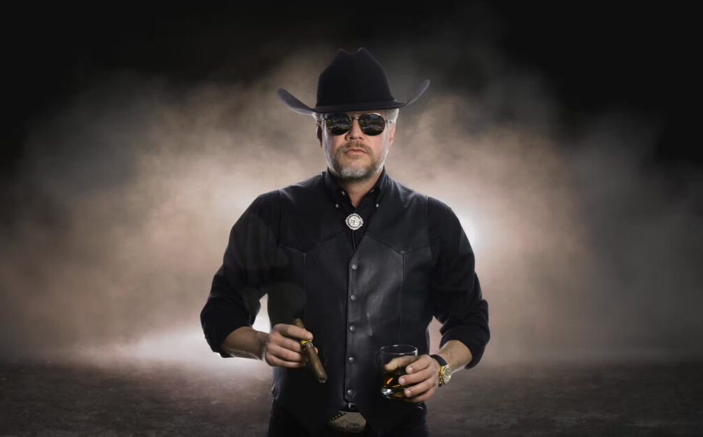 Man in black cowboy hat and vest, holding cigar and whiskey glass, wearing sunglasses. Dark background.