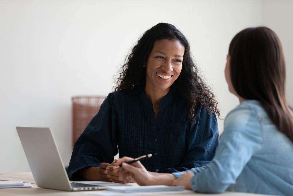 Woman smiles while speaking to another woman at a desk with a laptop.