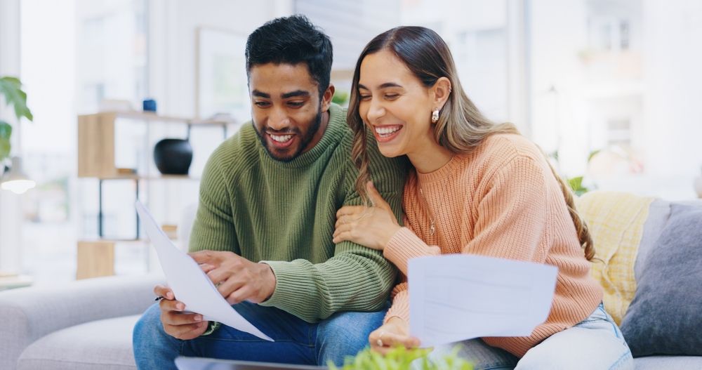 Couple smiles while reviewing papers on a couch in a well-lit living room.