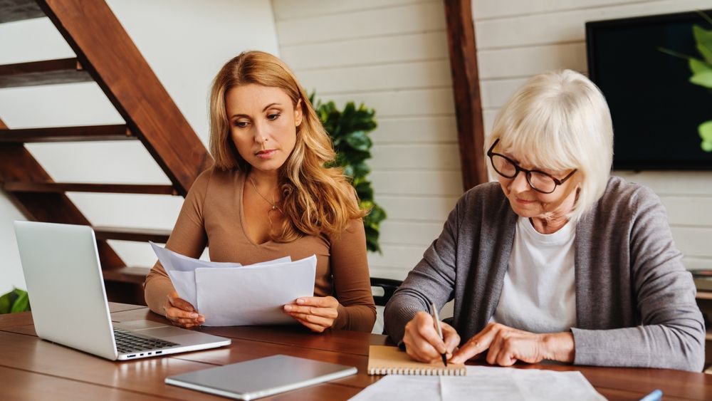 Woman and older person reviewing paperwork with a laptop and writing on a notepad.