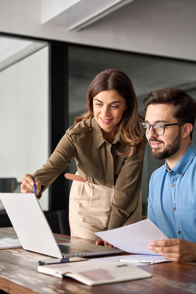 Woman pointing at laptop screen, man looks on, both smiling, paperwork and notebook on a wooden table.