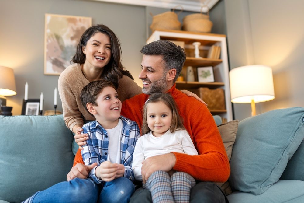 Family of four smiles together on a couch in a living room.