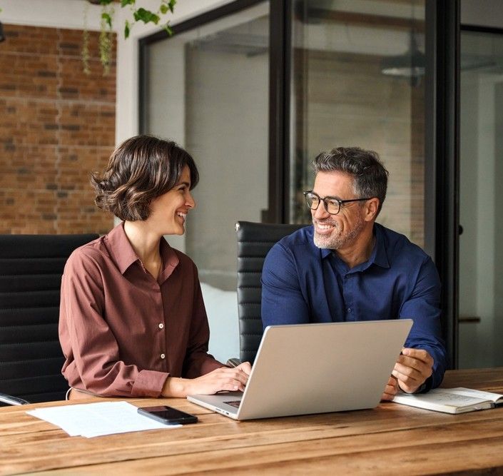Two people smiling at a laptop, sitting at a wooden desk in an office.