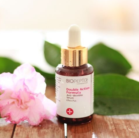 A bottle of double action formula anti wrinkle serum is sitting on a wooden table next to a pink flower.