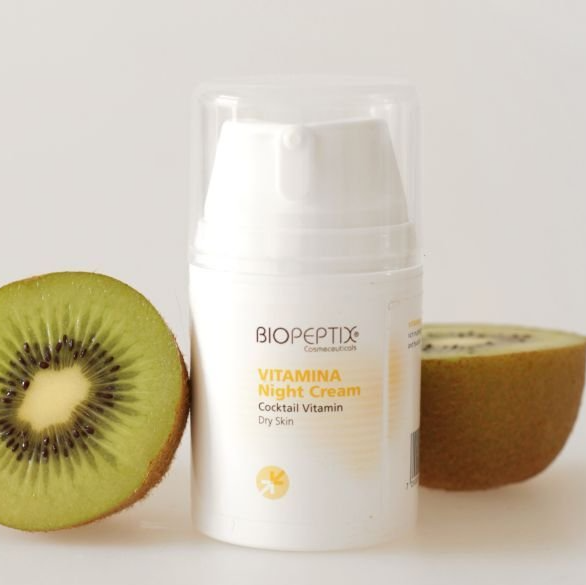 A bottle of biopeptide vitamina night cream next to a kiwi