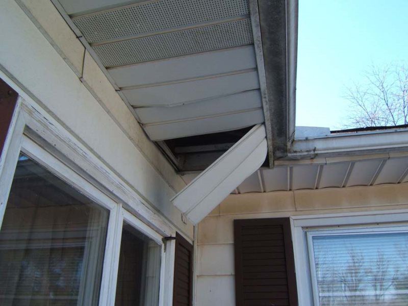 Damaged white siding on house eaves, with a section hanging down near a window and brown shutters.