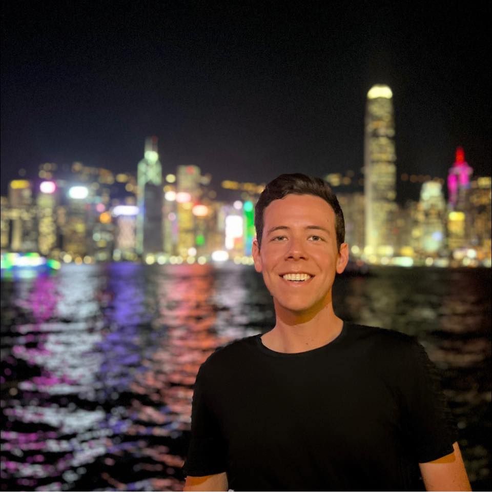 Man smiling in front of a nighttime cityscape with water in the foreground, possibly Hong Kong.