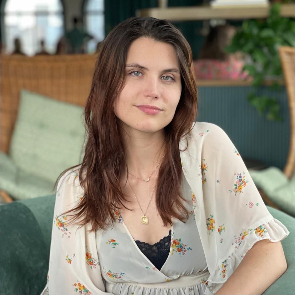 Woman with long brown hair and fair skin, wearing a floral top, smiles, in a well-lit setting.