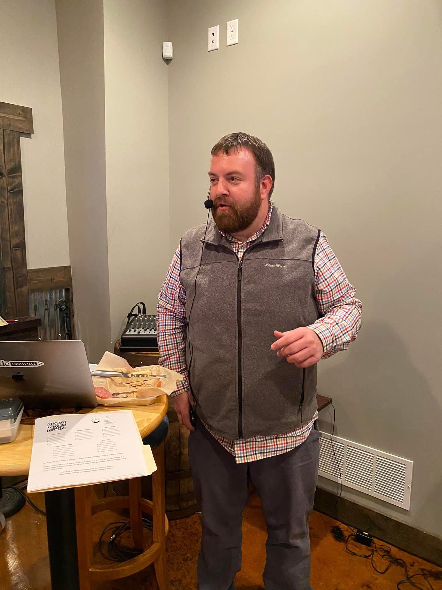 Man in vest speaking at an event, standing near table with laptop, microphone on, indoor setting.