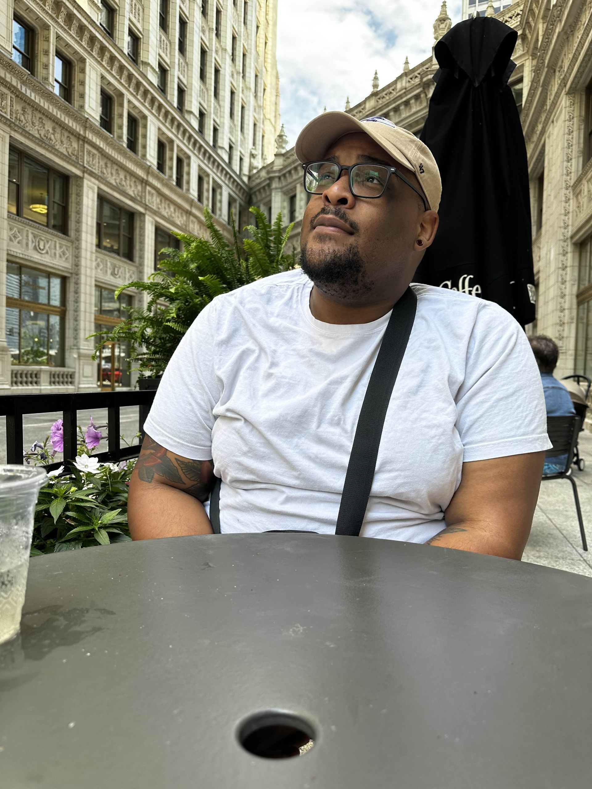 Man in a white t-shirt and cap looking up, sitting outside at a table in front of a building.