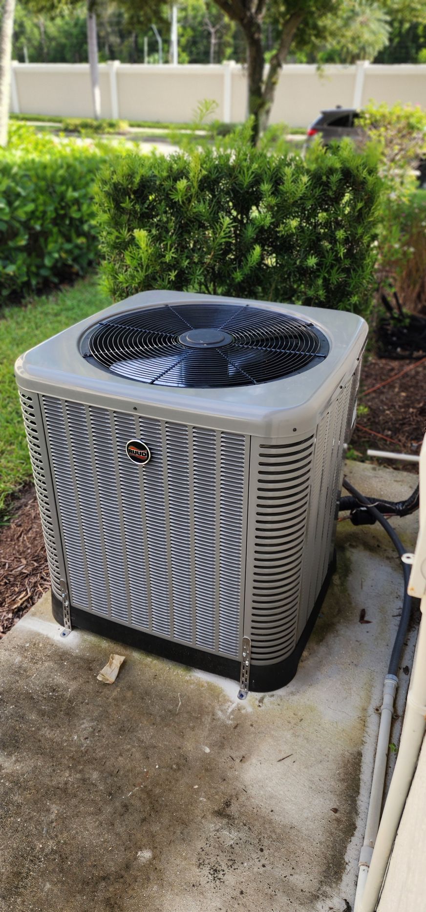 A gray air conditioner is sitting on a concrete patio next to a bush.