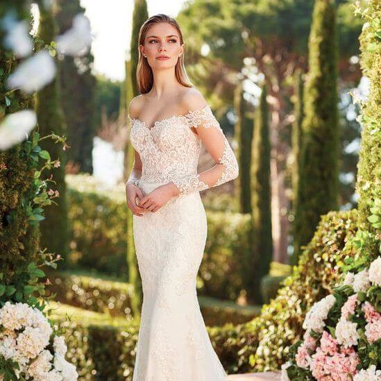 Woman in a white off-shoulder gown standing in a lush garden with floral arrangements