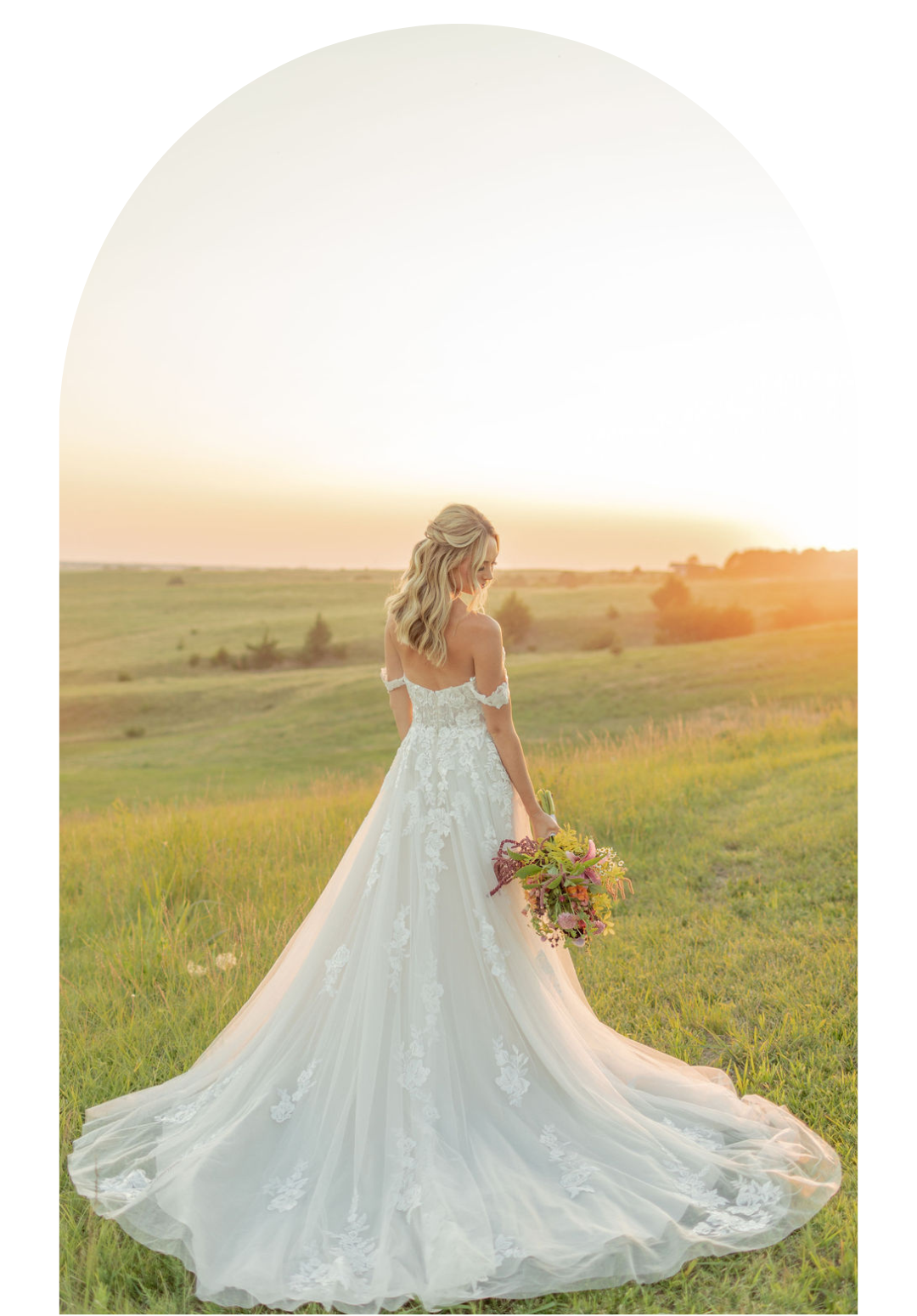 Bride in a white lace gown holding a bouquet in a sunlit field at sunset