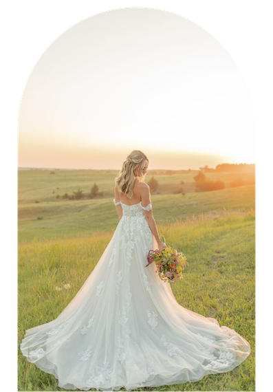 Bride in a white lace gown holding a bouquet in a sunlit field at sunset