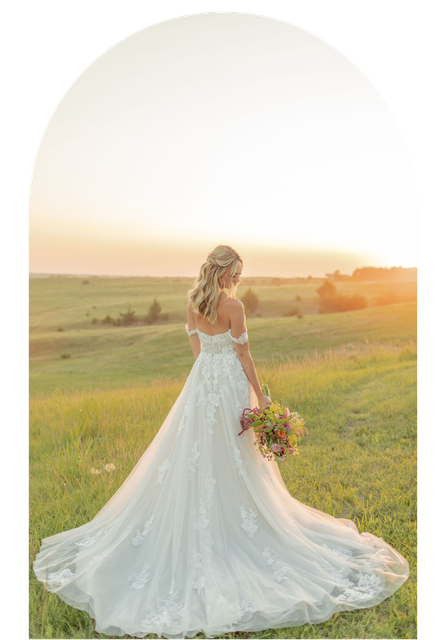Bride in a white lace gown holding a bouquet in a sunlit field at sunset
