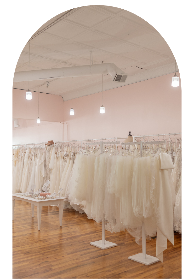 Rows of cream dresses on racks in a pastel pink boutique with arched ceiling and wood floor