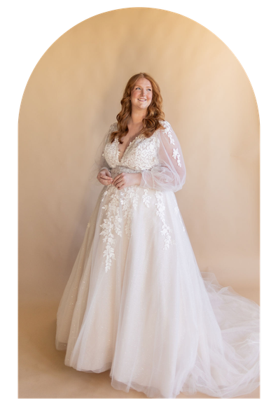 Woman in a white lace gown standing before a beige backdrop, holding her hands together and smiling.
