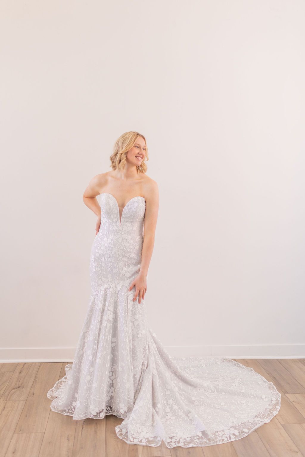 Bride in a white off-shoulder lace wedding gown standing with a flowing train against a plain wall