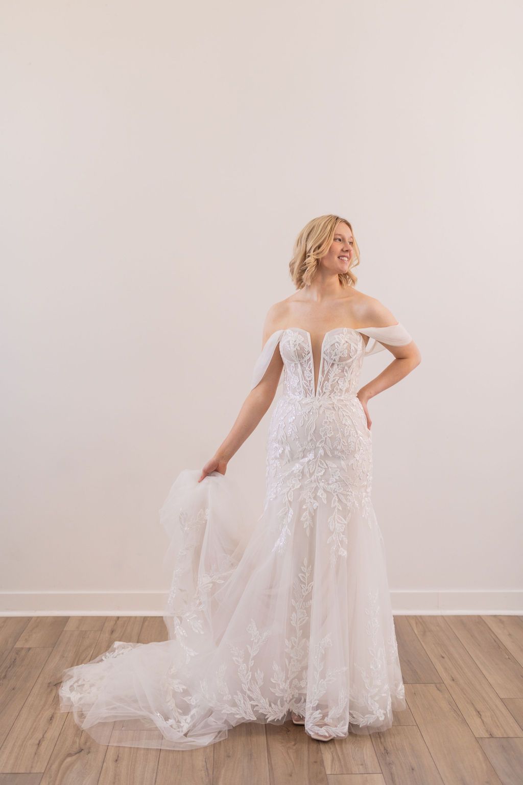 Bride in a white off-shoulder lace wedding gown standing with a flowing train against a plain wall