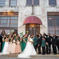 Wedding party posing in front of a brick building, bride in white and bridesmaids in green dresses.