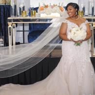 Bride in a white gown holding a bouquet on a decorated stage, with a long veil flowing behind her.