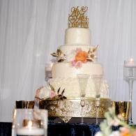 Three-tier white wedding cake with gold topper and flowers on a decorated table