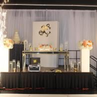 Elegant black-and-gold event dessert table with white drapes and floral decorations at a venue