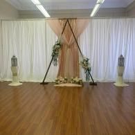 Decorated indoor event backdrop with white drapes, wooden arch, floral arrangements, and polished floor