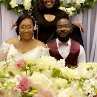 Wedding couple seated at a floral table, smiling before white and green floral decor