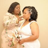 Two women in floral dresses share a tender moment, one adjusting the other’s attire while holding flowers.