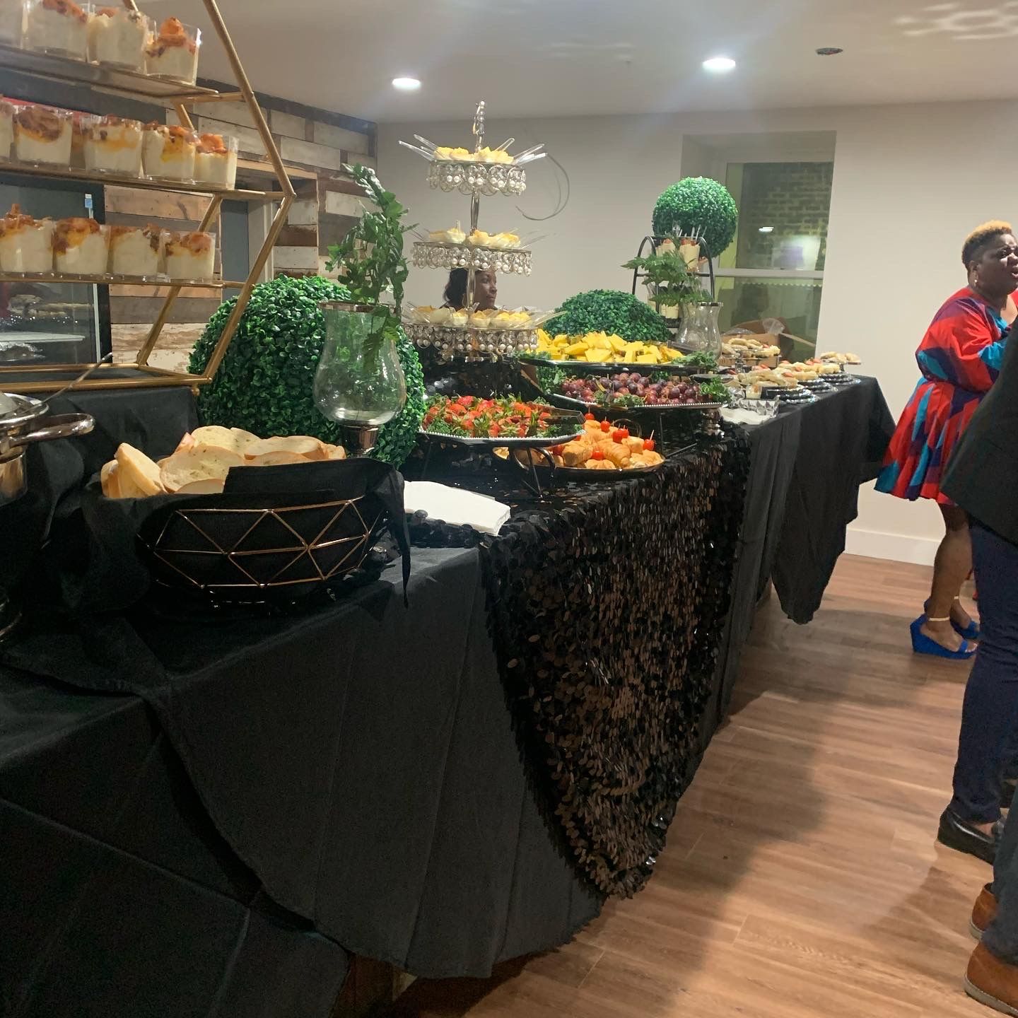 Buffet table with covered dishes and decorative greenery in a banquet hall, with a few guests nearby