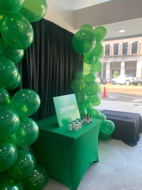 Green balloon-decorated booth with a black curtain backdrop and a “DEFENSE” sign on a table