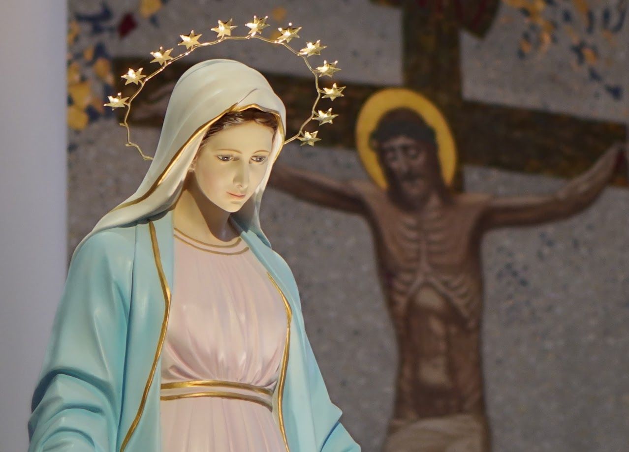 Statue of the Virgin Mary in blue and white, with a halo, in front of a crucifix in a church.