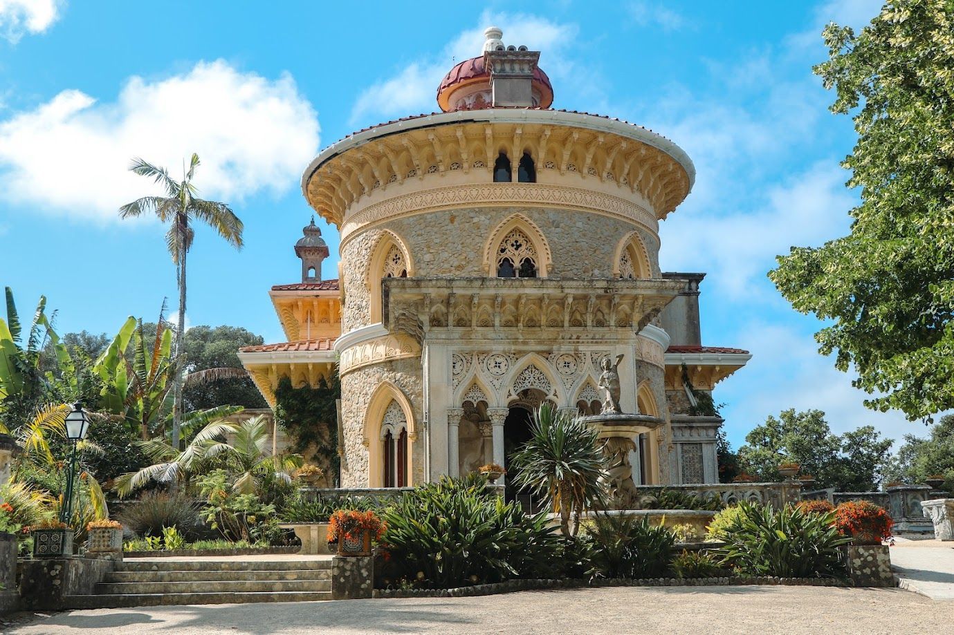 Ornate stone pavilion with a domed roof, surrounded by lush greenery under a bright blue sky.