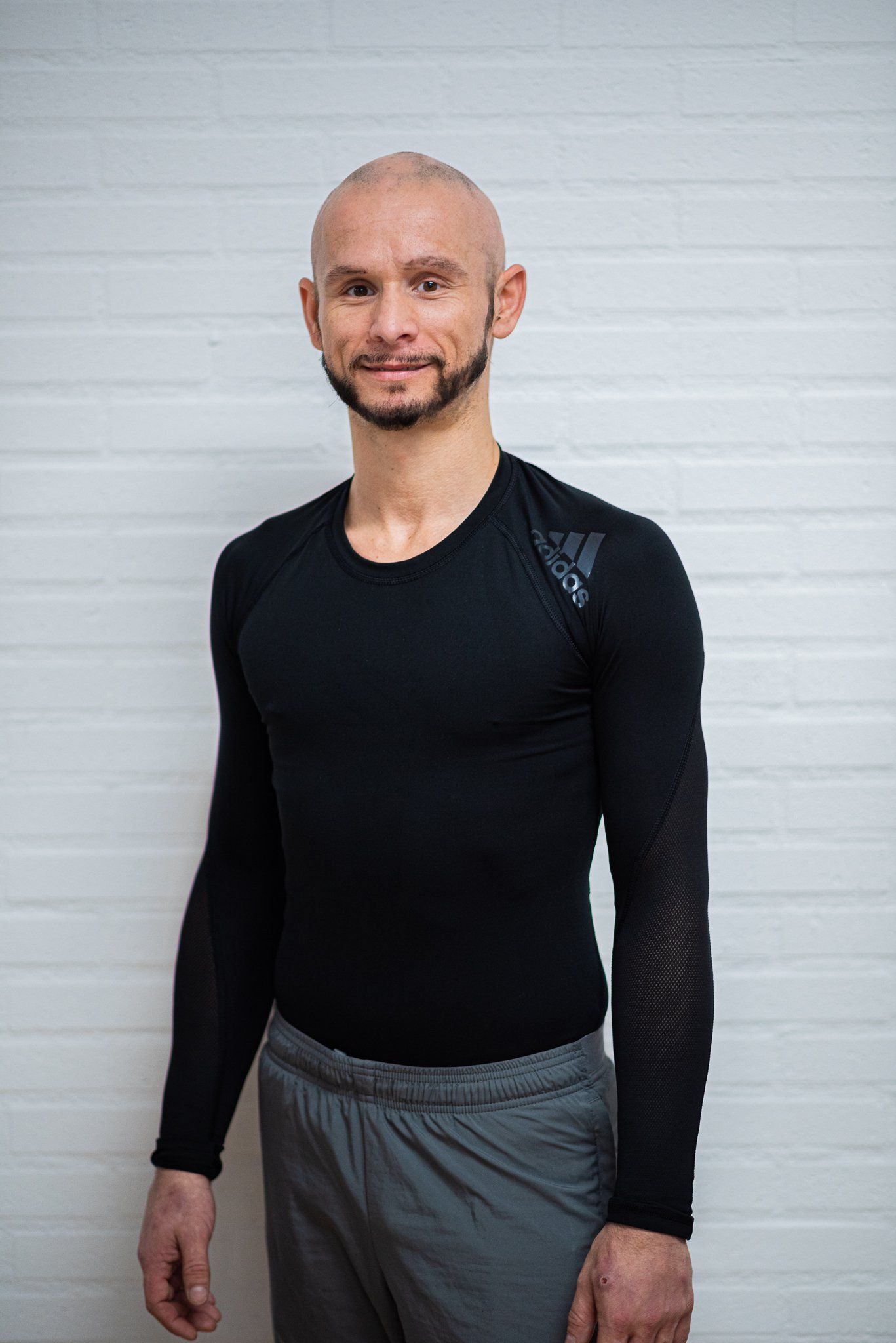 Een kale man met een baard, gekleed in een zwart shirt en een grijze broek, staat voor een witte bakstenen muur.