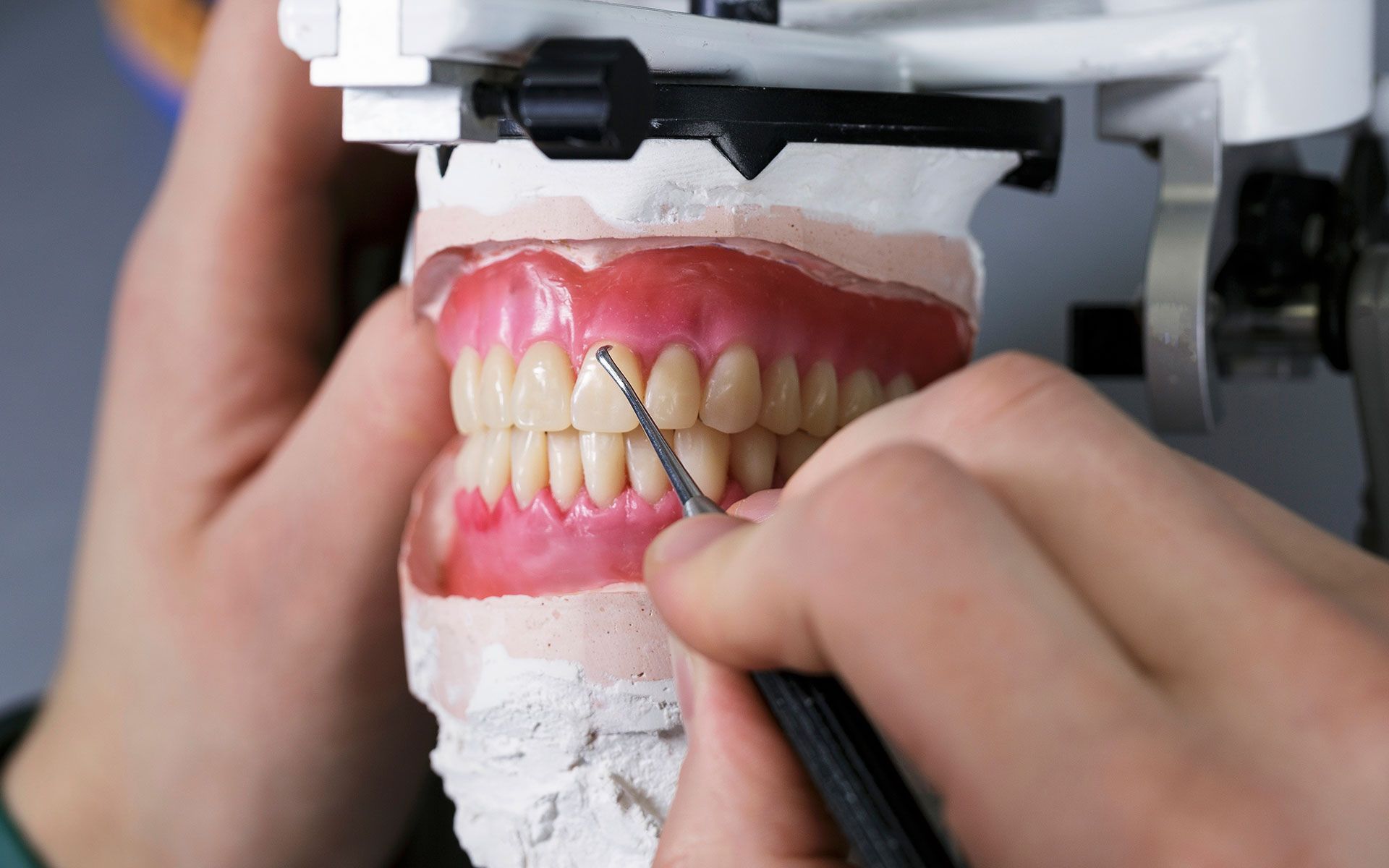 Hands working on a denture model with tools; the setting is a dental lab.