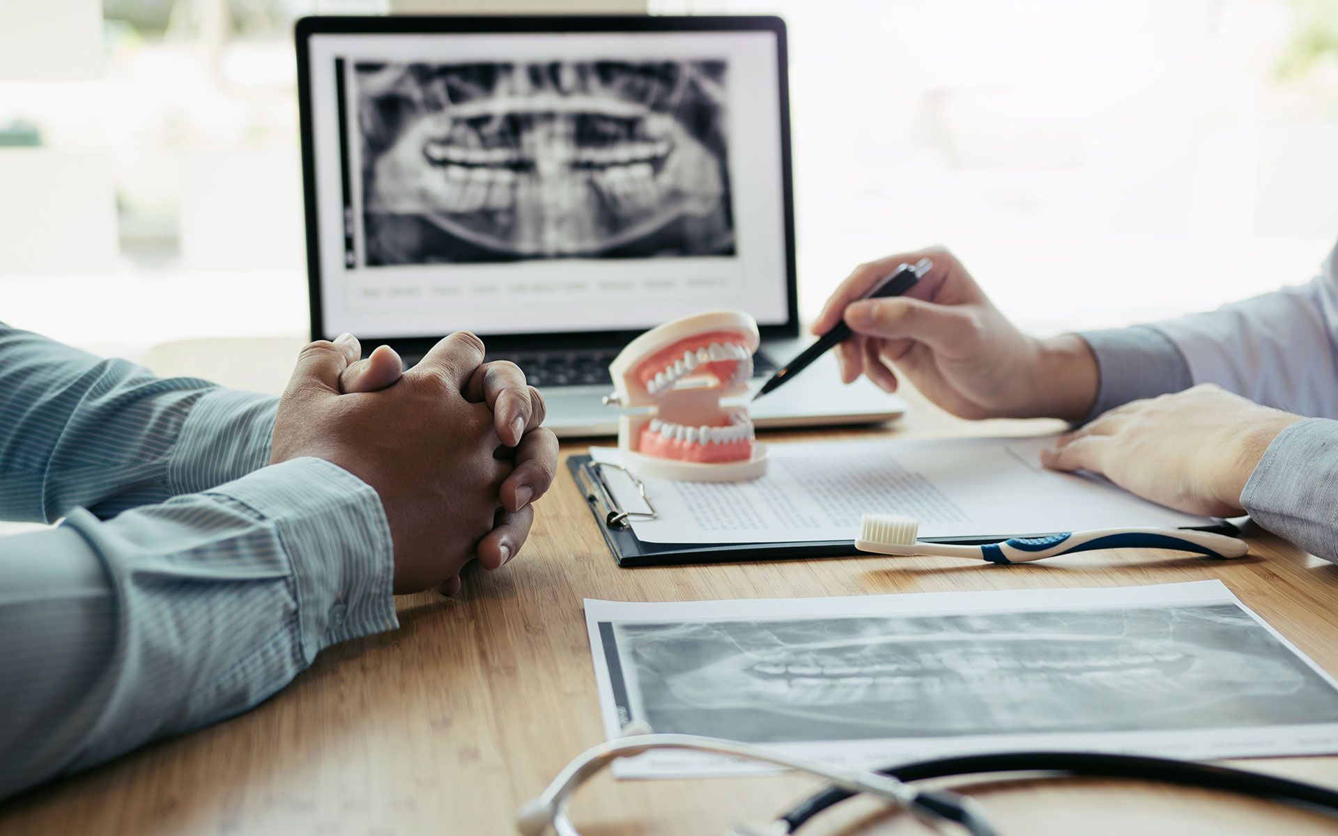Dentist pointing at X-ray on laptop, explaining to patient. Dental model and charts are on table.