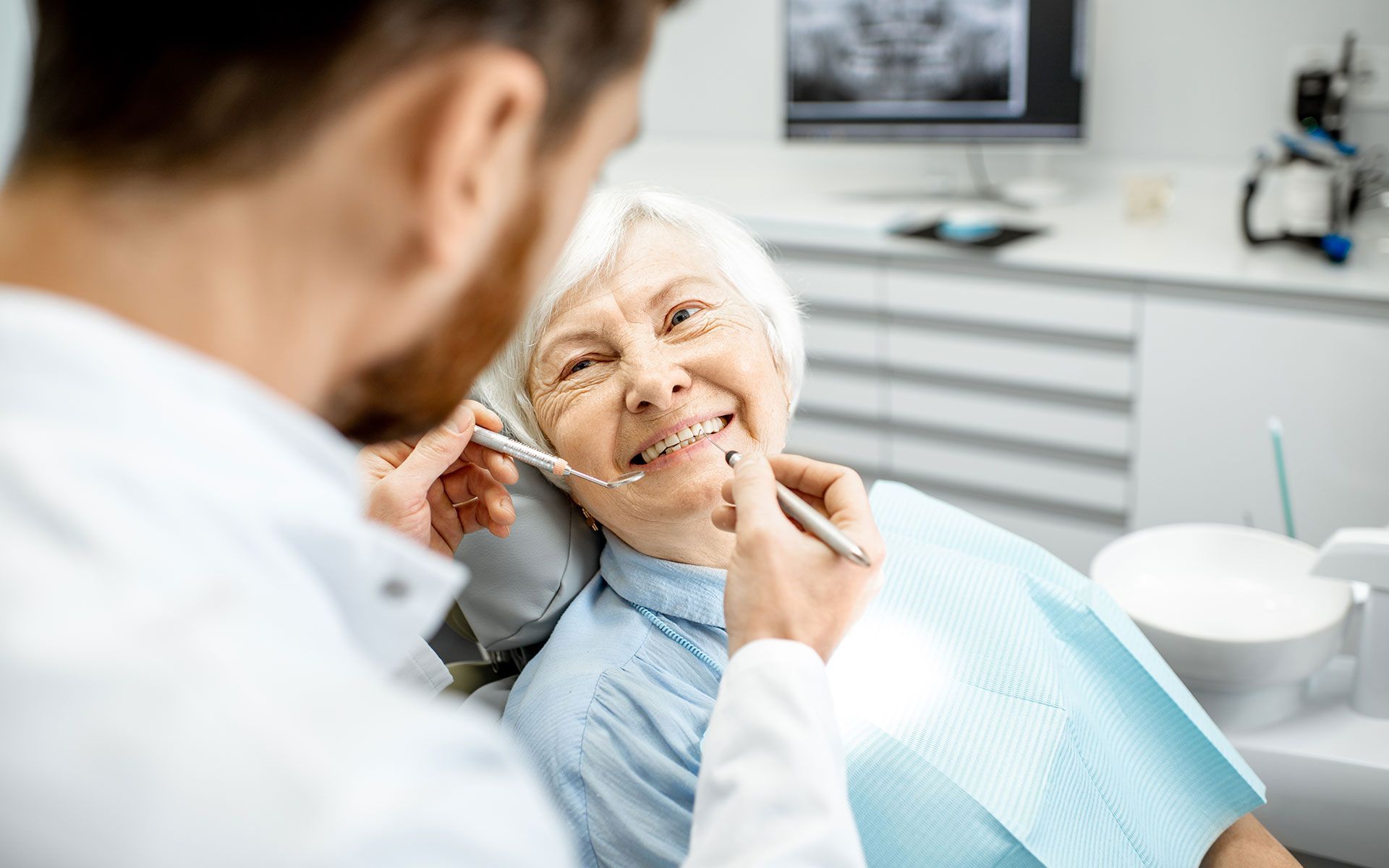 Dentist examining an elderly woman's teeth in a bright dental office.