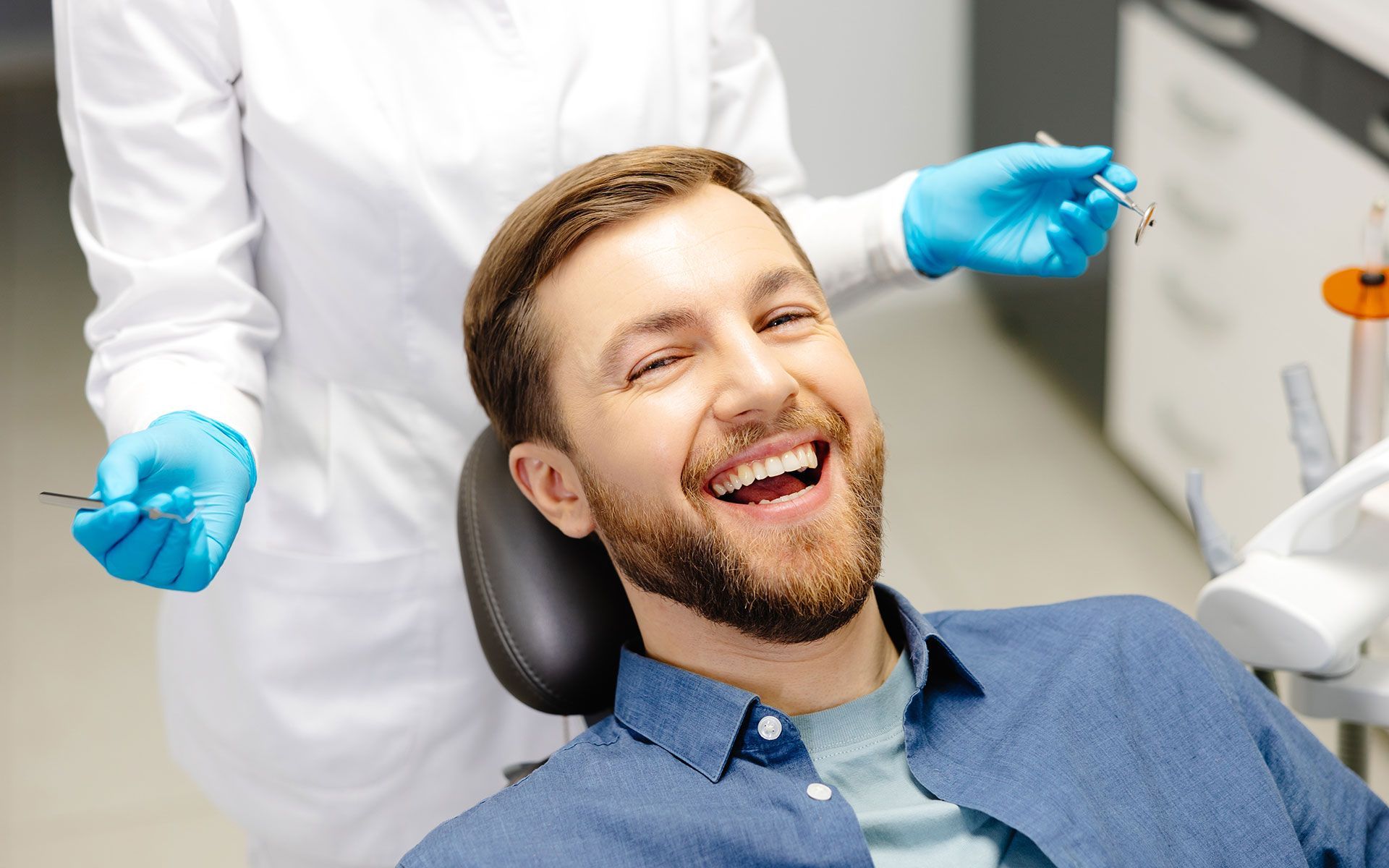 Man laughing in a dentist chair. Dentist in white coat and blue gloves holds tools.