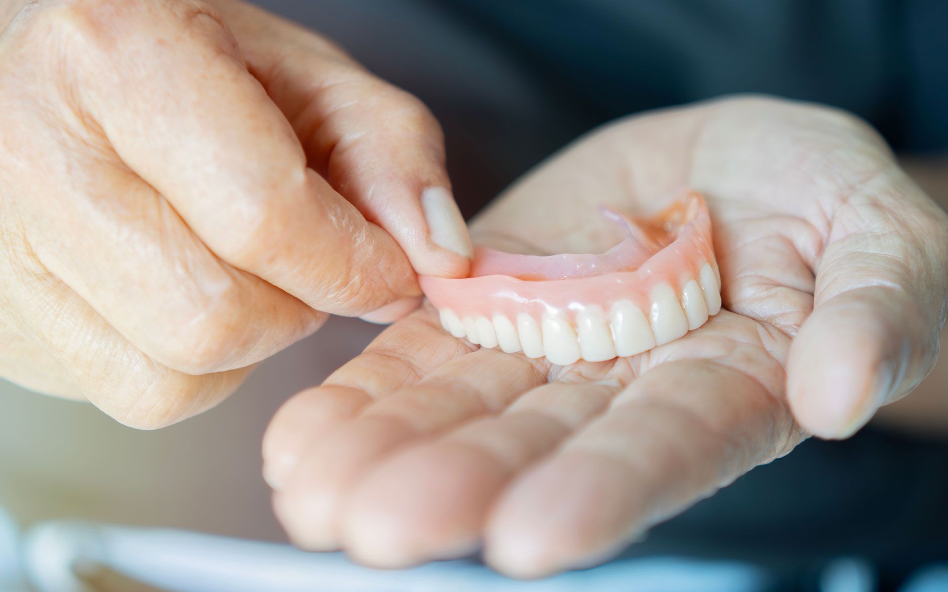 Person holding upper dentures in their palm. Pink base, white teeth, neutral skin tone.