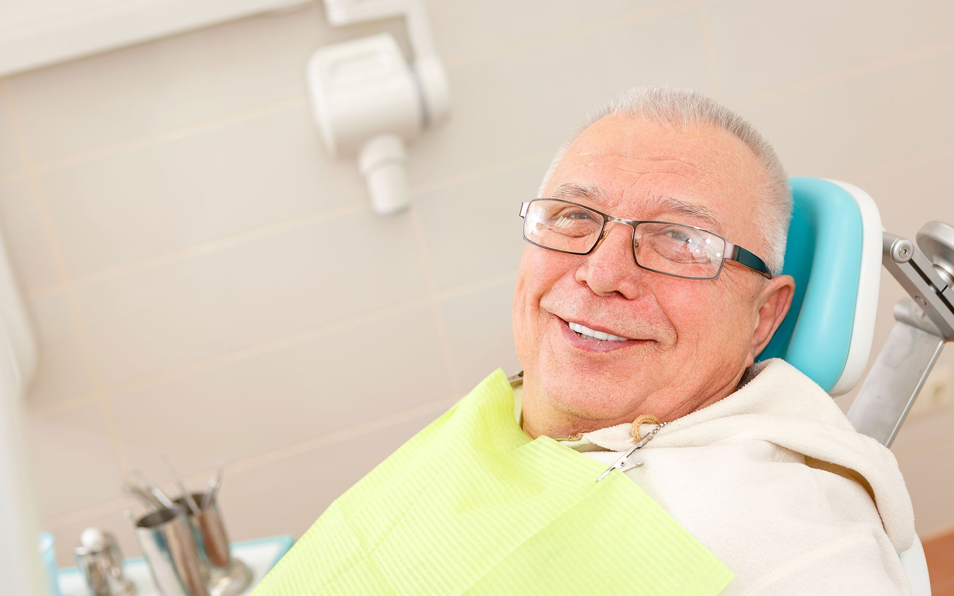 Smiling older man in dental chair, wearing glasses, with bib; dental setting.