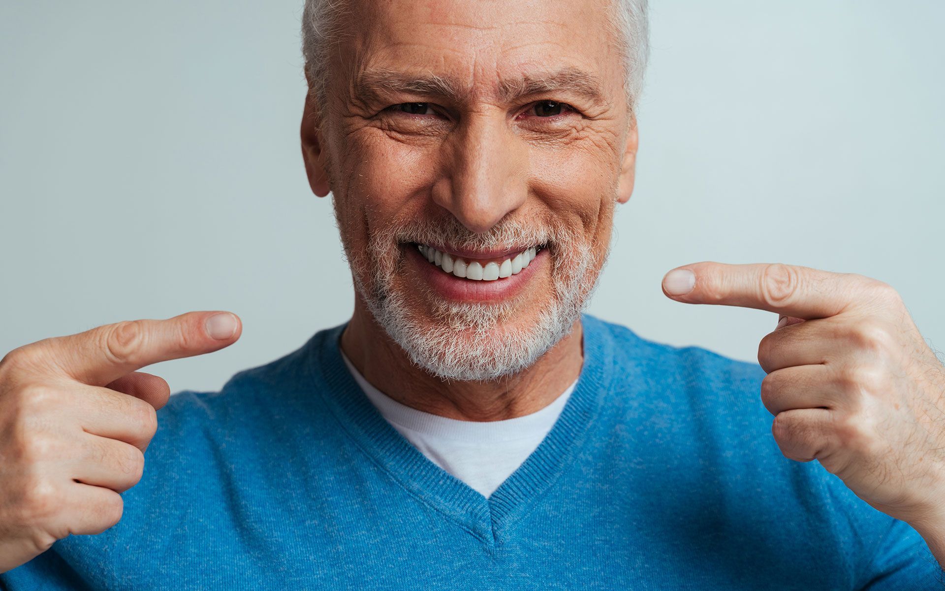 Smiling older man points to his perfect white teeth; light blue shirt, grey hair, light background.