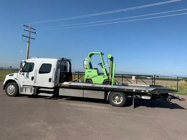 A white tow truck is towing a green forklift.