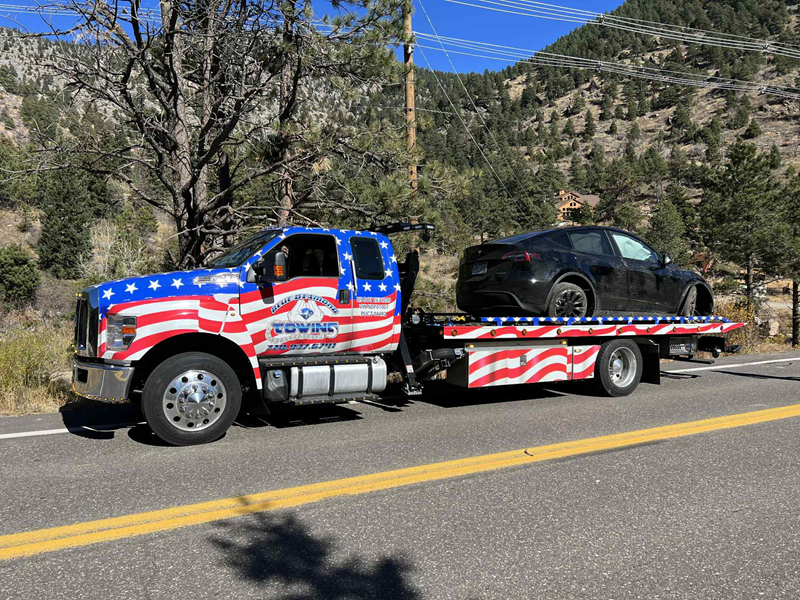 Tow truck with American flag design hauls a black car on a road, mountains in background.